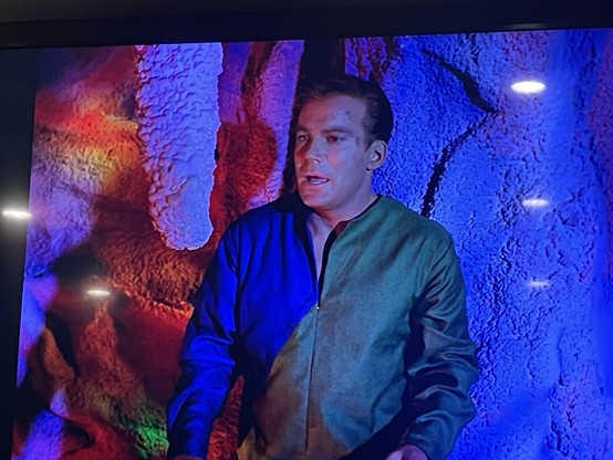 Captain Kirk stands in a blue and pink hued cavern. There is a large stalactite hanging in front of him. He is wearing a half green and half blue jumper.