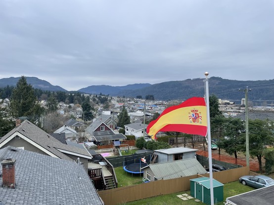  A spanish flag flies over a view of Port Alberni city with homes and trees and a cloudy sky with mountains in the distance.