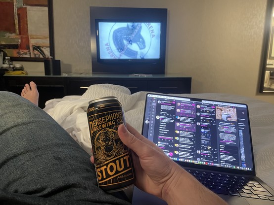 a picture from the hotel bed looking at the TV as the canucks logo shows with my laptop with mastodon up on the sreen and a Persephone stout 