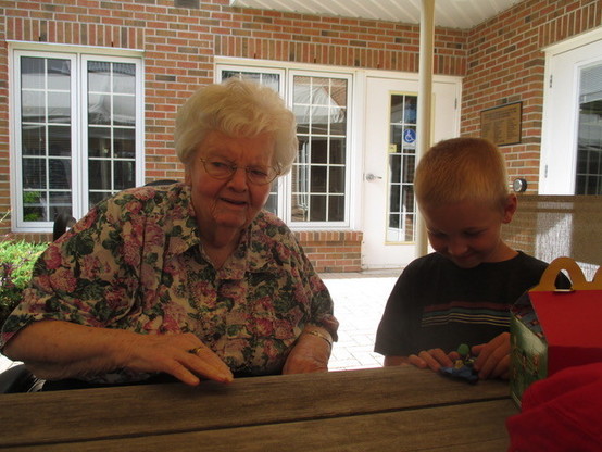 An elderly lady in a floral blouse sits up to a wood picnic table in the shade on a patio. She is in a wheelchair. A young boy stands beside her playing with a toy on the table while a young peers into a McDonalds happy meal box.