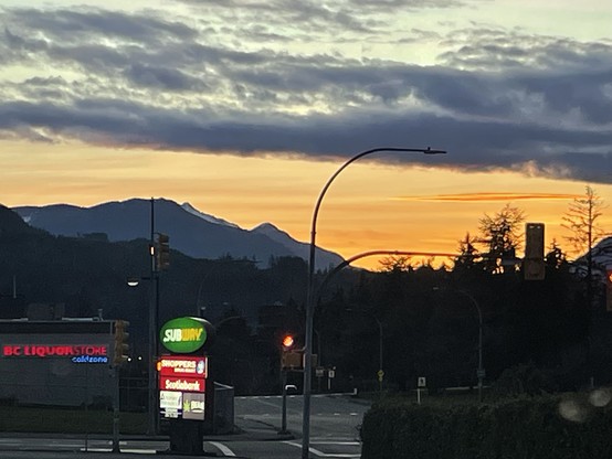 A sunset scene from a parking lot. In the foreground is a subway sign with some other stores underneath. And intersection. Above is a sky lit up orange by the sunset. There is multiple levels of mountain outlines. There are layers of clouds of different colors, orange, and yellow and gray.
