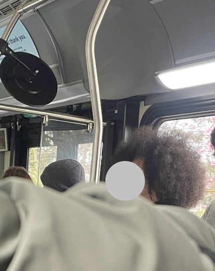 A person sits near a window on a bus. His face is digitally obscured for privacy. Only the side profile of his voluminous brown afro is visible.