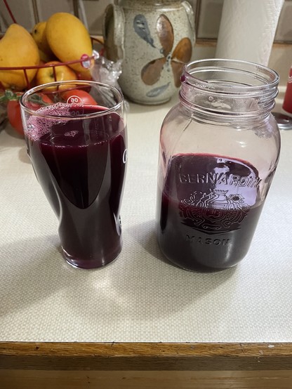 A glass and a jar stand on a white countertop. They are full and half full respectively of dark purple grape juice.