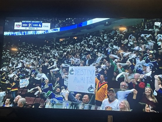 A jubilant Vancouver fan holds sign that says destiny awaits with two Vancouver Canucks logos on it. All around him are cheering and waving fans as the team  wins.