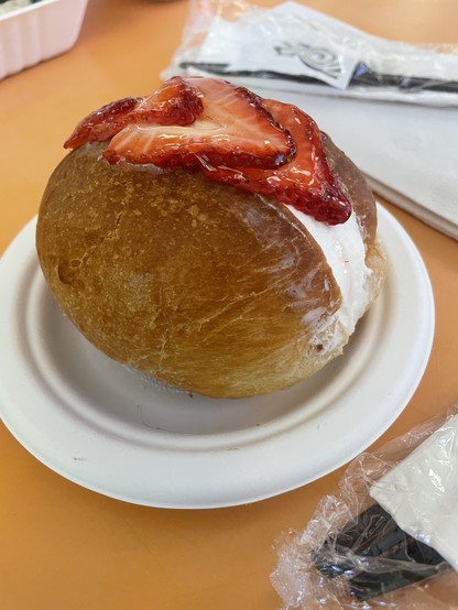 A bun sits on a small paper plate. It is large and round with a gap in the middle filled with cream and topped with slices of strawberries.