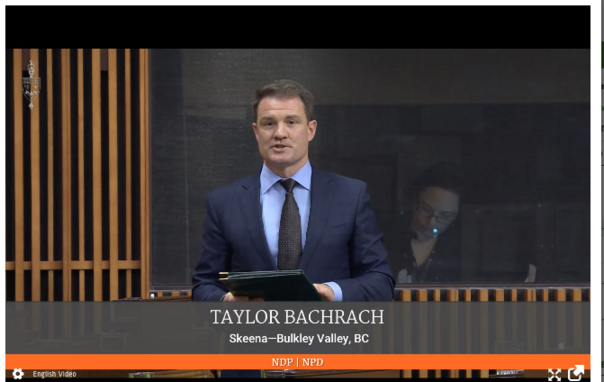 A screenshot of the video shows a man standing in a blue suit speaking. There is a Chiron on the bottom that says his name and constituency and party (in orange) as Taylor Bachrach Skeena-Bulkley Valley BC NDP/NPD