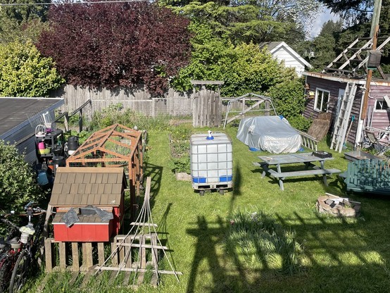 The cart with the tank on top is sitting in the middle of a large grassy backyard between the garden boxes and chicken coop on the left and the shed and small car under tarp on the right.