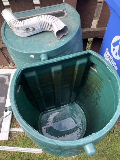 two green flat backed water barrels. One has had its top cut out revealing the empty depths inside. The other one behind it has a angled piece of white downspout lying on top of it. 