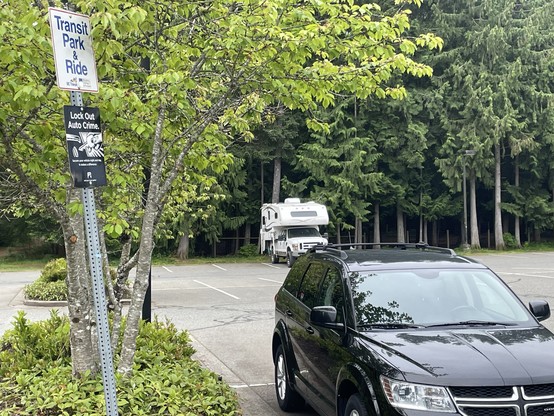A black Dogde Journey sits parked in a stall beside a small parking lot island that has trees and a tall metal sign post with two signs One saying "Transit Park &Ride and another "Lock Out Auto Crime"