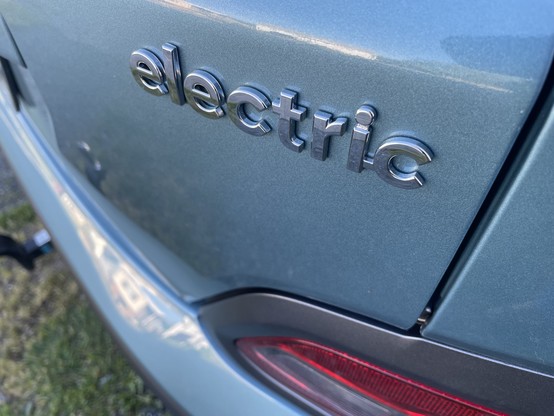 A picture of the back passenger side corner of a Hyundai Kona electric. It is a blue color. Centred in the picture sort of skewed is the word electric.