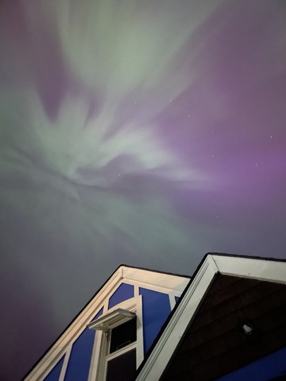 A picture looking up toward the night sky. In the bottom portion is the double peak of our house with its bright white trim and bright blue paint. There is a tall window with one window swung open hinged from the top.

Above the house is an electric purple sky with a green splash of colour from the aurora. Behind, the colour the stars are visible.