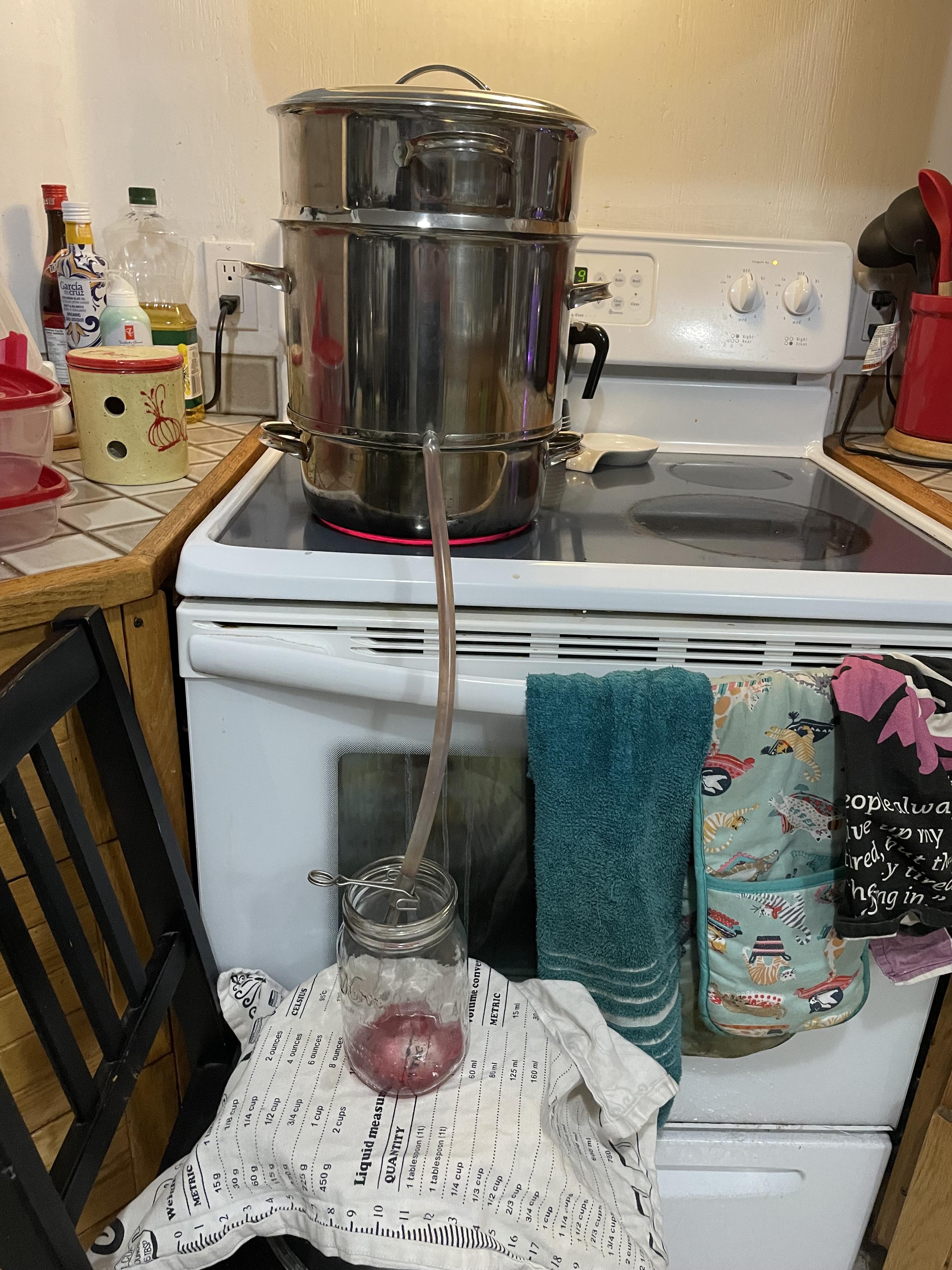 this picture shows the three level steamer sitting high on top of the stove top with a plastic tube coming from the bottom that is emptying juice into a jar that is standing on top of the pot, which is standing on top of a chair