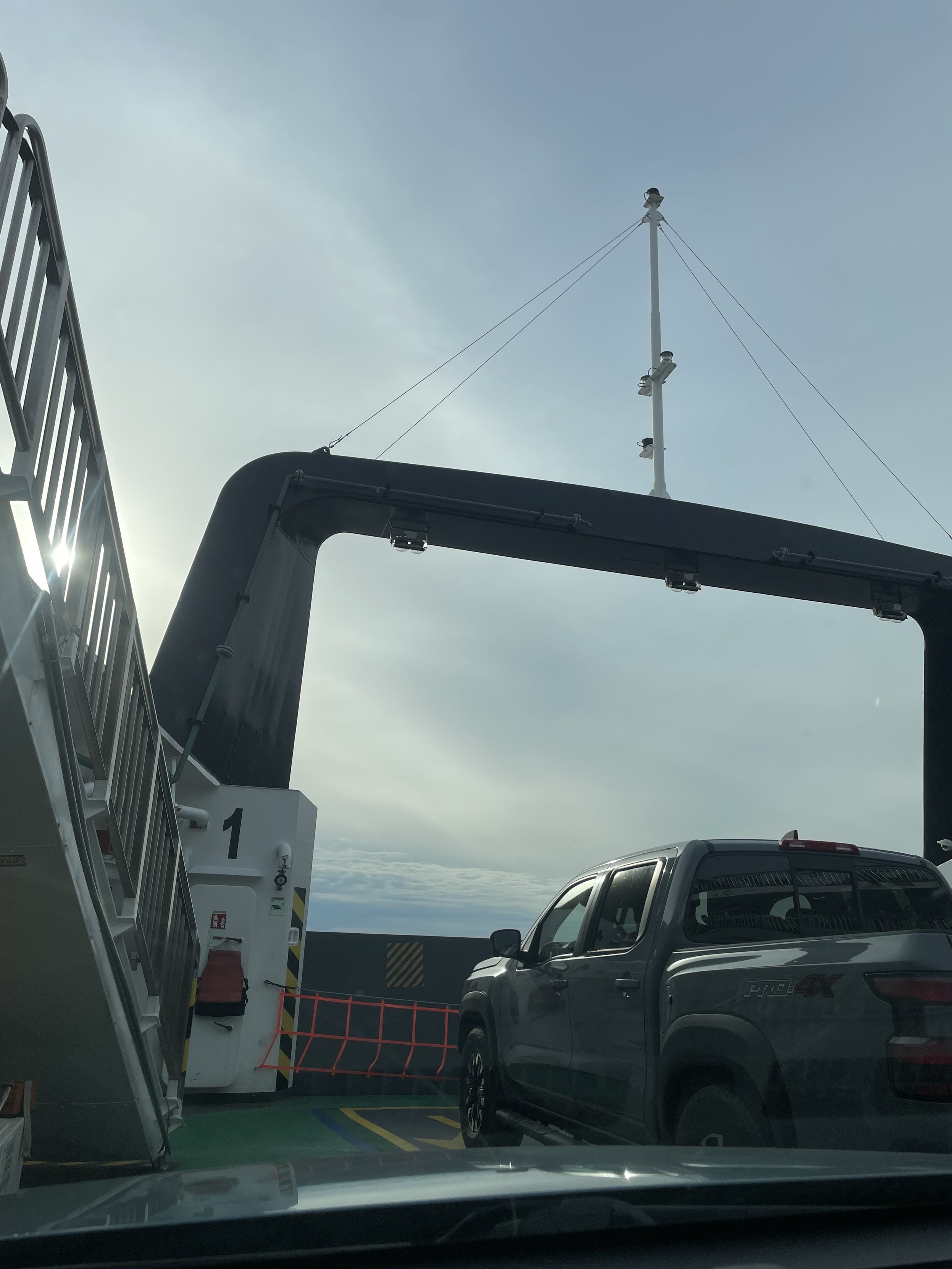 A view from the lower deck of the ferry. I am the first car but there is a truck to my right in front. A small stair case stretched up toward me on the left. There is a large black square bracket thing thay kind of looms over the front