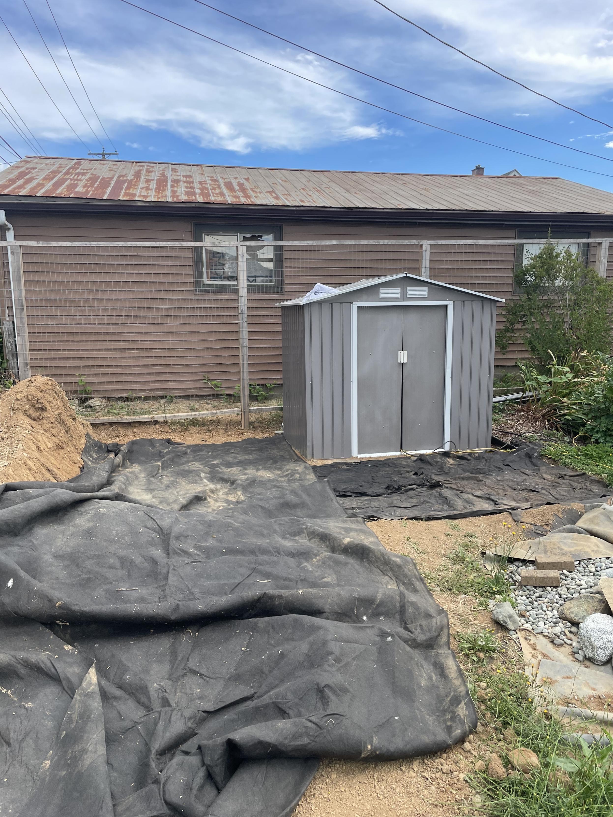 A huge piece of black cloth lays out in front of into the side of the shed. There are other pieces in front of the shed. I will be cutting a huge piece down to fit the space that isn't covered by the small pieces.